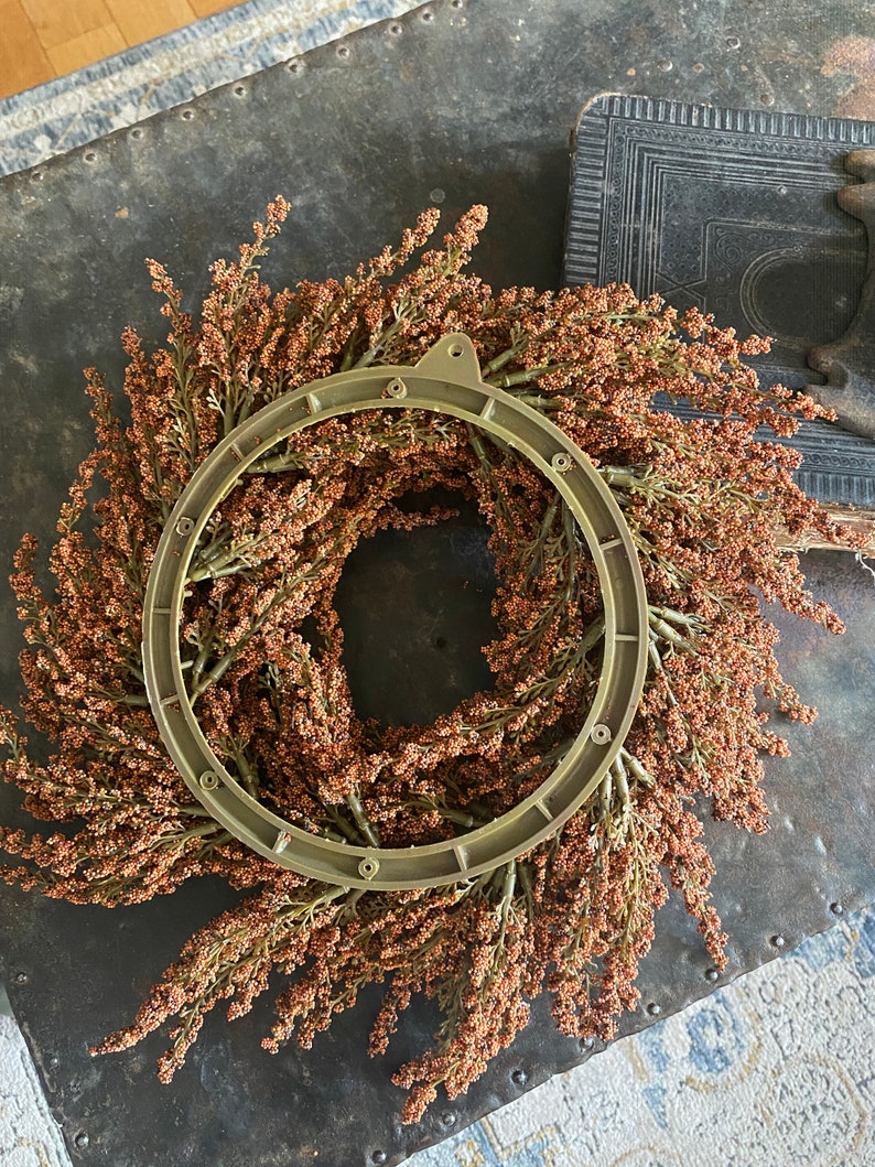 May include: A decorative wreath featuring a circular metal frame with a ring of dried, brown-toned floral elements. The wreath is set against a dark, textured surface, suggesting a rustic or vintage aesthetic. The overall design evokes a natural, autumnal theme.
