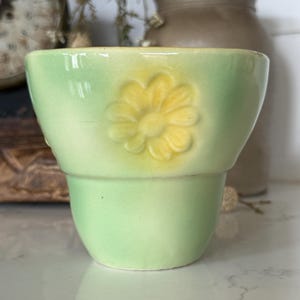 May include: A light green ceramic planter with a yellow flower design. The planter has a two-tiered design and a glossy finish. The flower is embossed on the front of the planter. The background includes a clock and a brown vase.