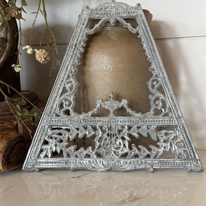 May include: A triangular silver-toned metal trivet featuring an intricate openwork design with ornate scrollwork, leaf motifs, and a central lyre emblem. The piece has a weathered, textured finish and a wide base with small feet for stability.
