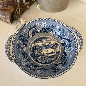 May include: A round ceramic bowl featuring a blue and white transferware pattern. The center depicts a detailed landscape with buildings and boats on water, while the rim is decorated with intricate oak leaves, acorns, and a geometric border.
