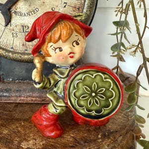 May include: A ceramic figurine of a child dressed in a red hat and boots, and a green outfit. The figurine is holding a drum with a green floral design. The background includes a vintage clock and decorative greenery.