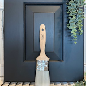 May include: A close-up of a paintbrush with a wooden handle and a silver ferrule. The brush is labeled "Royal & Langnickel 1" and is resting on a white wooden surface.