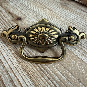 May include: Antique brass drawer pull with a floral center design and curved handle. The pull has ornate detailing on the sides and a slightly aged appearance, suggesting vintage or retro style.