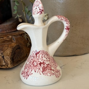 May include: A cream-colored ceramic cruet featuring a red floral transferware pattern. The vessel has a wide base, a curved handle, a pouring spout, and a matching pointed stopper. The surface displays a fine crackle glaze finish throughout.