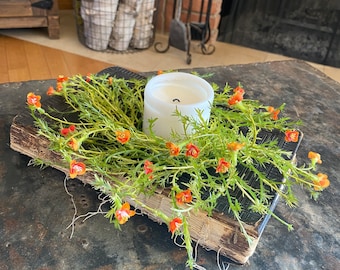 May include: A white candle sits in the center of a wreath of orange flowers on top of an old, worn book. The book is lying on a dark gray metal surface.