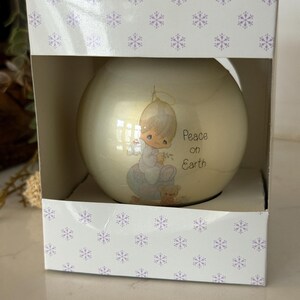 May include: A round, off-white Christmas ornament featuring a cherubic figure with the words "Peace on Earth." The ornament is inside a white box decorated with purple snowflakes.