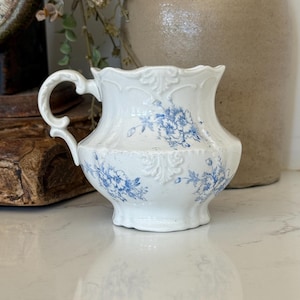 May include: A white ceramic creamer pitcher featuring a scalloped rim, an ornate handle, and embossed detailing. The surface is decorated with a blue floral pattern, showcasing delicate blossoms and foliage across the rounded body of the vessel.