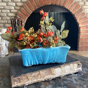 May include: A blue ceramic planter with a wavy rim, filled with a faux floral arrangement of orange and red flowers and green leaves. The planter is sitting on top of a vintage book with a worn leather cover.