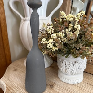 May include: A gray wooden muddler with a rounded top and a long, slender handle. The muddler is displayed on a wooden surface, with a white vase and a floral arrangement in the background. The muddler is a kitchen tool.