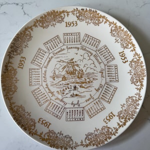 May include: A vintage white plate with gold accents and a 1953 calendar design. The plate features a windmill scene in the center, surrounded by monthly calendars and floral patterns. The year "1953" is repeated around the rim.