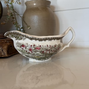 May include: A cream-colored ceramic gravy boat featuring a detailed transferware pattern of a rural landscape with red-roofed houses, green trees, and flowering branches, finished with a dark brown decorative border along the top rim and handle.