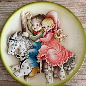 May include: A decorative ceramic plate featuring a carousel scene with two children riding a spotted horse. The plate has a light green border and the name "Sarah Kay" is printed on the right side.