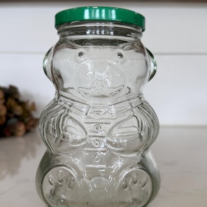 May include: A clear glass bear-shaped jar with a green lid. The jar has embossed details, including a bear face, paws, and a button-down shirt design. The jar is designed to hold honey or other food items.