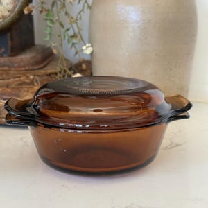 May include: A round, amber-colored glass casserole dish with a matching lid. The dish features two integrated side handles and a smooth, translucent finish. The lid has a textured, circular pattern around the center, providing a clear view of the interior.