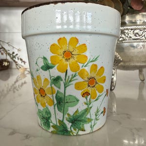 May include: A white ceramic flower pot with a speckled finish and a brown rim. The pot is decorated with yellow daisy-like flowers, green leaves, and stems. A decorative item for home decor.