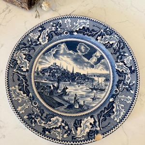 May include: A round ceramic plate featuring a blue and white transferware pattern. The center depicts a harbor scene with ships, buildings, and figures. The border is decorated with detailed oak leaves and acorns on a fine mesh background.