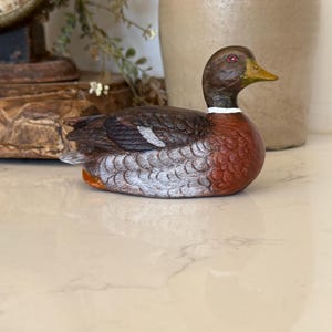 May include: A decorative duck figurine featuring a brown head, a yellow beak, and a white ring around the neck. The body displays a mix of dark brown, reddish-brown, and grey feathers with detailed texture, capturing the bird in a resting pose.