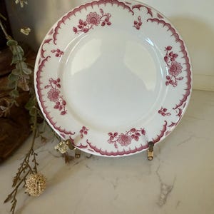 May include: White ceramic plate with a maroon floral pattern around the rim. The plate is displayed on a gold-colored stand. The floral design includes various flower and leaf motifs. The plate is a vintage style.