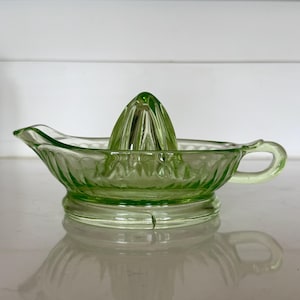 May include: A translucent light green glass citrus juicer featuring a central reamer cone, a wide bowl with a scalloped rim, a small pouring spout on one side, and a circular handle on the opposite side, all crafted with a pressed glass pattern.