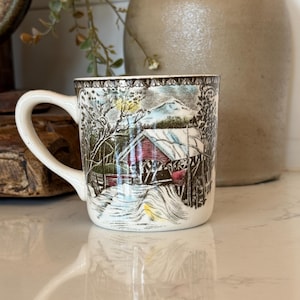 May include: A cream-colored ceramic mug featuring a detailed, multi-colored illustration of a red covered bridge in a snowy winter landscape. The scene includes bare trees, a mountain in the background, and a decorative brown border along the top rim.