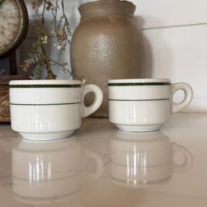 May include: Two white ceramic cups with a rounded handle and two thin horizontal dark green stripes encircling the upper portion of the body. The cups feature a straight-sided design with a slightly recessed base, reflecting on a smooth surface.