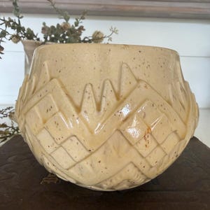 May include: A beige ceramic bowl with a geometric pattern. The bowl has a textured surface and a wide opening. The design features a series of zigzags and diamond shapes. The bowl is sitting on a dark brown surface.