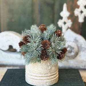 May include: A decorative arrangement of faux pine needles and pine cones in a cream-colored, textured vase. The arrangement features blue-green needles and brown pine cones, creating a natural, seasonal display. The vase has horizontal grooves.