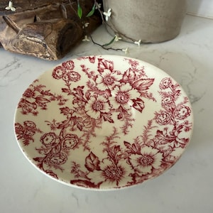 May include: A round ceramic saucer featuring a cream-colored base with a detailed dark red floral pattern. The design consists of various flowers, leaves, and intricate botanical elements covering the entire surface of the dish in a repeating style.