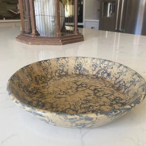 May include: A round, antique-style bowl with a beige base and a blue, marbled pattern. The bowl has a slightly raised rim and is likely made of ceramic or stoneware. The bowl is sitting on a white countertop.
