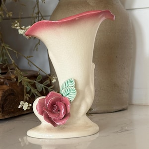 May include: A cream-colored ceramic vase with a flared, pink-rimmed opening. The base features a sculpted, three-dimensional dark pink rose blossom with two light green leaves attached to the side of the vase body, which has a subtle ribbed texture.