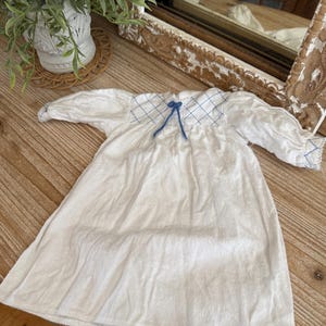 May include: White baby gown with long sleeves. The neckline features a blue grid pattern and a blue bow. The gown is made of soft fabric and is laying on a wooden surface. The gown is a vintage style.