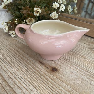 May include: A pink ceramic creamer with a white interior and a handle. The creamer is sitting on a wooden surface.