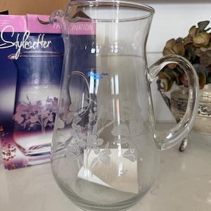May include: A clear glass pitcher with a handle and a floral etched design. The pitcher is next to a box with the word "StyleSetter" on it. The pitcher is on a white surface.