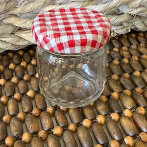 May include: A clear glass jar with a red and white checkered lid. The jar is empty and sits on a wooden beaded surface. The lid is a classic design, often used for preserving jams and jellies.
