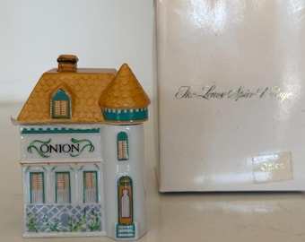 May include: A porcelain spice jar shaped like a house with a yellow roof and a turret. The front features the word ONION in black text, green trim, and a blue lattice pattern at the base. It is accompanied by a white box labeled The Lenox Spice Village.