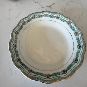 May include: A round ceramic bowl with a scalloped edge, featuring a white center and a decorative border of green leafy garlands and small brown flowers. The rim is accented with a thin gold-colored trim, and the surface shows a fine crackle glaze.