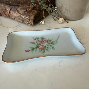 May include: A rectangular porcelain tray with a slightly curved, wavy edge and a gold-painted rim. The center of the white tray features a delicate hand-painted floral motif consisting of pink rose blossoms, buds, and green leaves on thin stems.