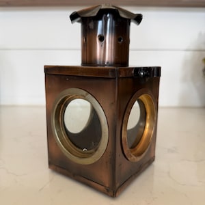 May include: A copper-toned metal lantern with a square base and a cylindrical top. The lantern has circular glass windows and a decorative top. The metal has a slightly aged appearance, suggesting a vintage or antique style.