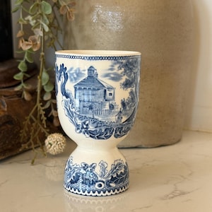 May include: A cream-colored ceramic egg cup featuring a blue transferware pattern. The design depicts a landscape with a building, trees, and a person, surrounded by decorative oak leaves and acorns around the base and rim.