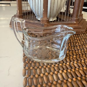 May include: A clear glass creamer with a handle and a spout, featuring a floral design. The creamer sits on a wooden beaded mat. In the background is a decorative birdcage and a white ceramic pitcher.