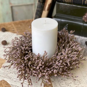 May include: A white pillar candle sits in the center of a wreath made of dried lavender sprigs. The wreath is on a wooden table with old papers and books in the background.