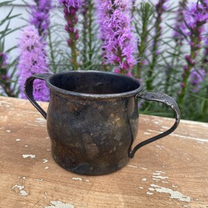 May include: A silver cup with two handles. The cup has a tarnished finish and is sitting on a wooden surface.