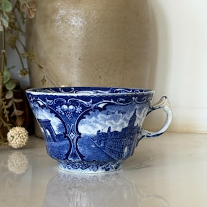 May include: A blue and white ceramic teacup featuring a detailed transferware pattern of architectural landmarks, including a building labeled City Hall, with ornate decorative borders and a curved handle.