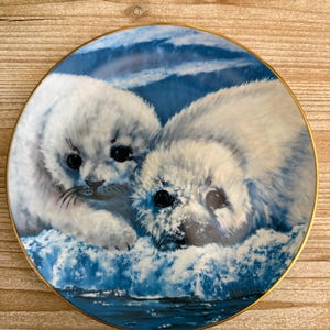 May include: A decorative plate featuring two white seal pups resting on a blue and white ice floe. The plate has a gold rim and a detailed illustration of the seals. The plate is likely for display or decorative purposes.