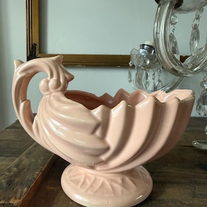 May include: A vintage, light pink ceramic planter with a scalloped rim and a pedestal base. The planter has a curved handle and a textured, swirled design. The base features a leaf pattern. The planter is displayed on a wooden surface.
