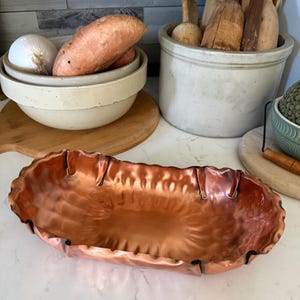 May include: A hammered copper serving dish with a ruffled edge. The oblong bowl has a shiny, reflective surface. The dish is empty and sits on a white countertop. Other kitchen items are in the background, including bowls of produce and wooden utensils.