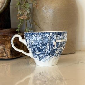 May include: A white ceramic teacup featuring a detailed blue transferware pattern depicting a horse-drawn carriage with the text TALLY-HO on the side. The cup has a curved handle and a scalloped rim, showcasing a historical scene in blue ink.