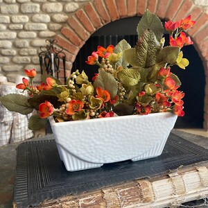May include: A white ceramic planter with a textured surface and four legs. The planter is filled with a bouquet of orange and white flowers with green leaves.
