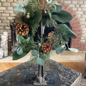 May include: A green and brown artificial wreath with pine cones and greenery is displayed on a vintage book stand. The stand is made of black metal and has a decorative design. The wreath is a festive decoration for the holiday season.