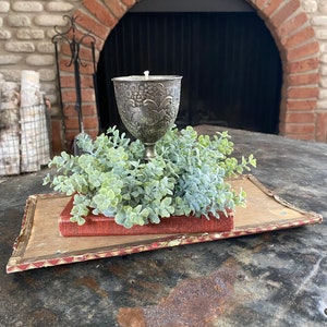 May include: A silver goblet candle holder with an ornate design sits on a red book surrounded by a wreath of green foliage. The candle holder is in front of a brick fireplace.
